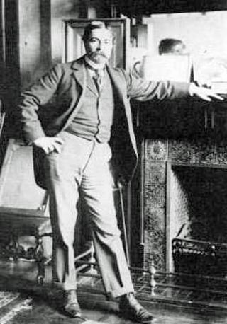 Photo of Childe Hassam posed standing by a fireplace,  date and location unknown.